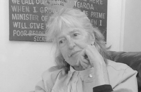Black and white photo of an old woman sitting with her left hand near her face, a poster hangs behind her on the wall.