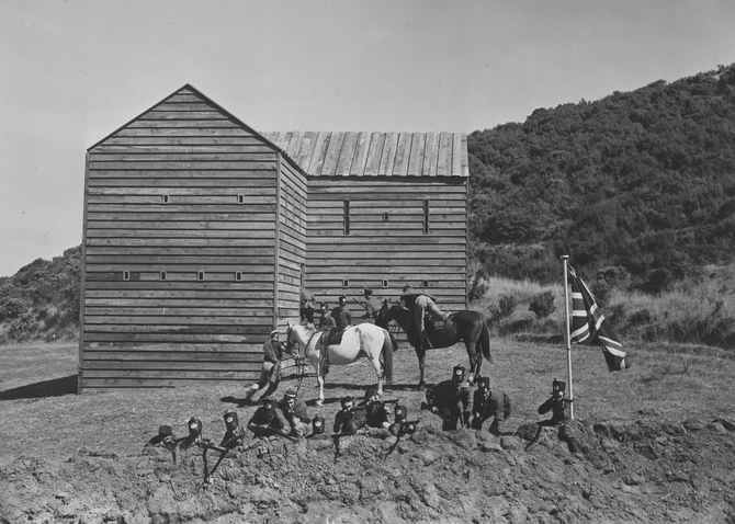 Soldiers prepare to fire rifles alongside flag. Wooden fortress behind them and two horses with riders.