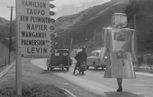 black and white photo of a road sign on the left, a car, a scooter and a person wearing a robot constume walking on the road 