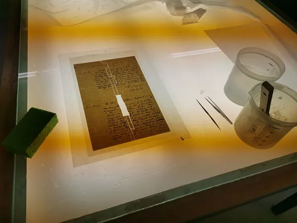 A piece of paper with pieces missing at the centre sits on an illuminated table. Next to the paper is a pair of tweezers and a pot of paste.h a brush inside it.