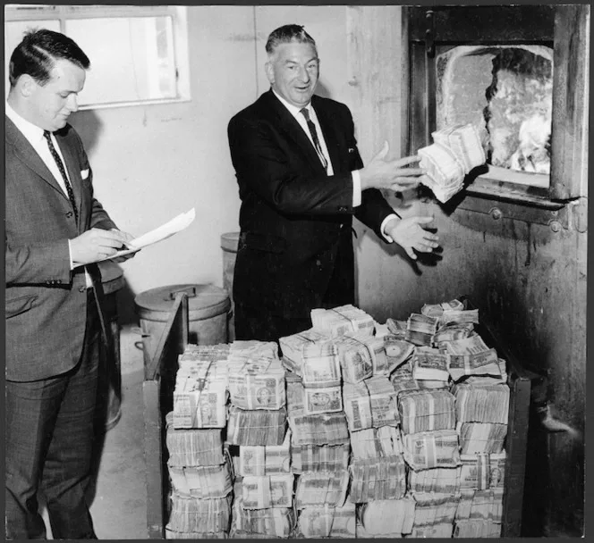 Black and white photo of two men wearing suits with a big pile of bank notes, one of them is throwing the notes in a  furnace