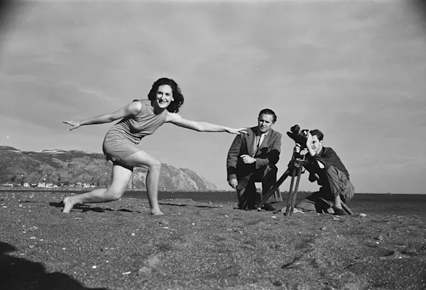 Three people filming at a beach. Closest is a woman dancing. Behind her two men crouch behind a camera.