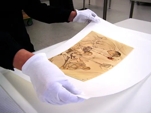 Photograph of a small paper artwork sitting on a larger, white piece of paper. A person wearing gloves holds the larger paper at the corners.