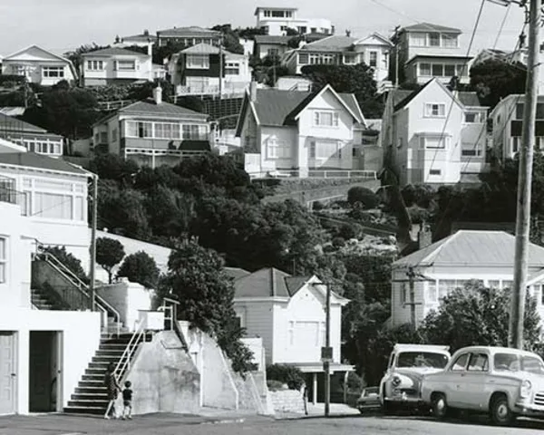 Rows of houses sit on a hillside. In the foreground there are parked cars and people walking on the footpath.