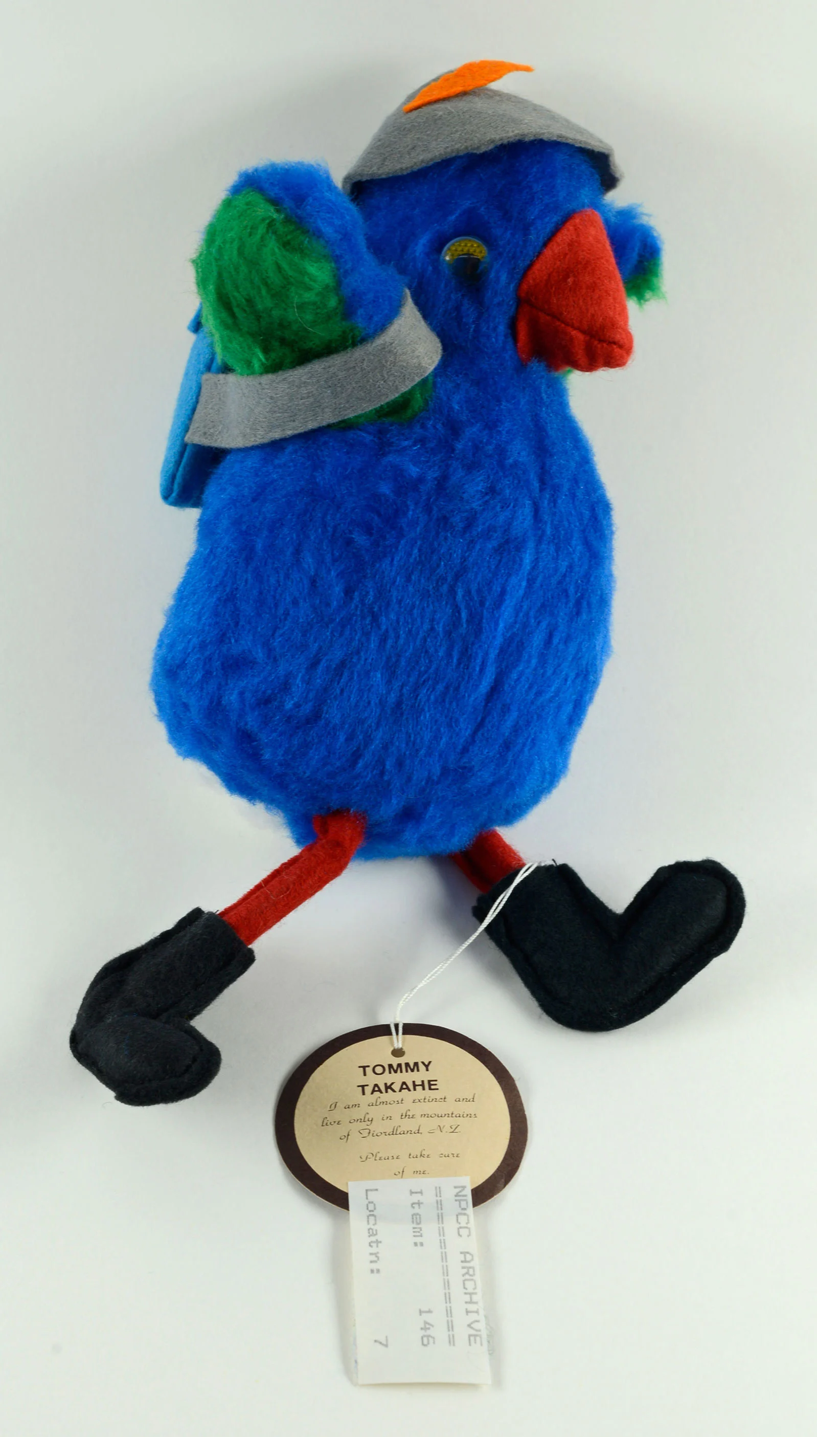A soft toy of a blue bird with red beak wearing a hat, black shoes and carrying a backpack