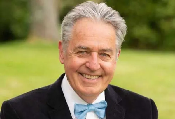 A smiling man with grey hair is wearing a black cost and blue bow tie.