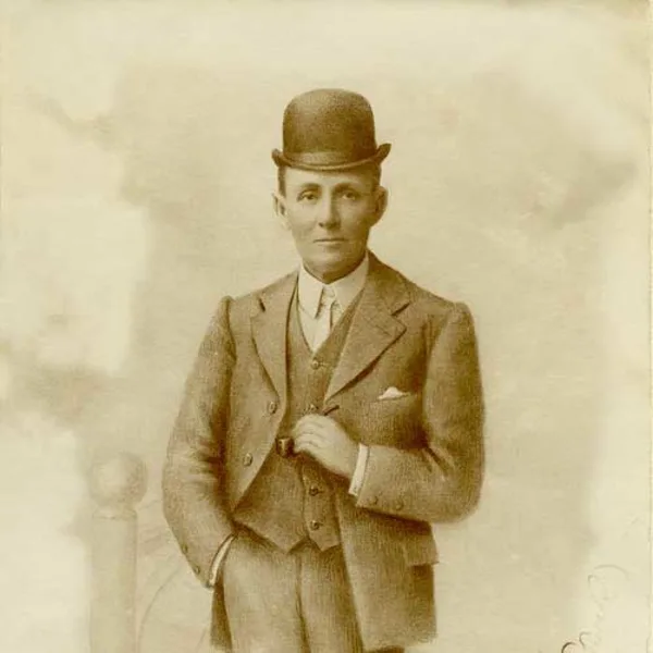 An old photo of a man wearing a suit and a hat standing in front of a pale yellow background, holding a pipe in his left hand and right hand in his pant's pocket