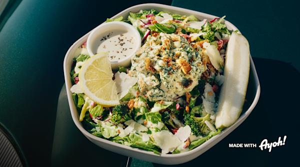 A hexagon white bowl of chopped salad on a dark green table: romaine, shredded red cabbage, roasted broccoli, and shaved parmesan, topped with a scoop of creamy chicken salad sprinkled with fresh dill and crispy breadcrumbs. A lemon wedge, a whole pickle spear, and a small cup of creamy dressing with cracked black pepper sit on the side.