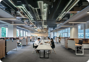 Tre professionisti lavorano a un tavolo condiviso in un moderno ufficio open space caratterizzato da un soffitto con grafiche luminose.