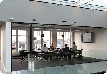 Four people in a meeting inside a glass-walled conference room with a view of the city.