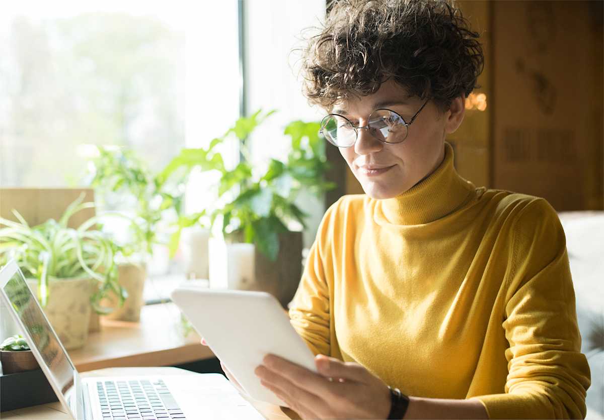 Person sitting at desk next to laptops and plants, looking at tablet