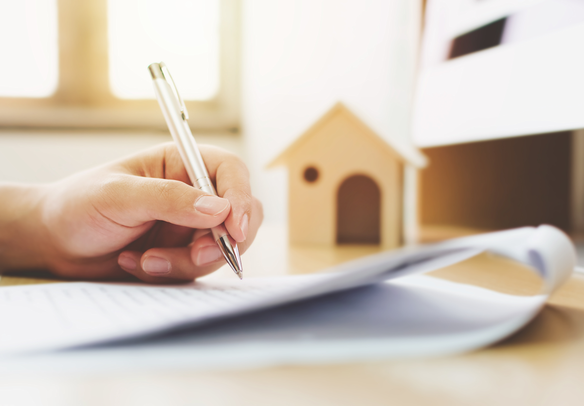 Hand with pen and paper next to a small wooden house