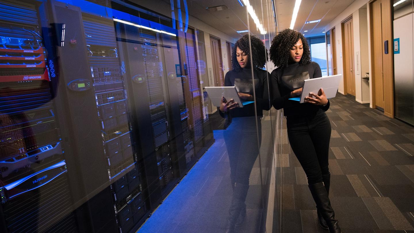 Image of a woman leaning on a glass wall with servers behind it holding a laptop.