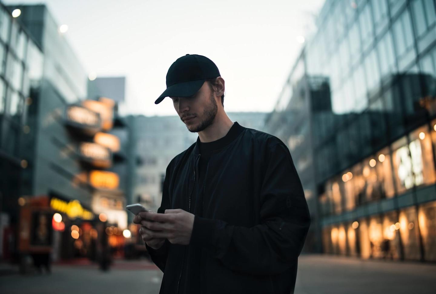 A man in a cap looking at his phone with blurred buildings behind him
