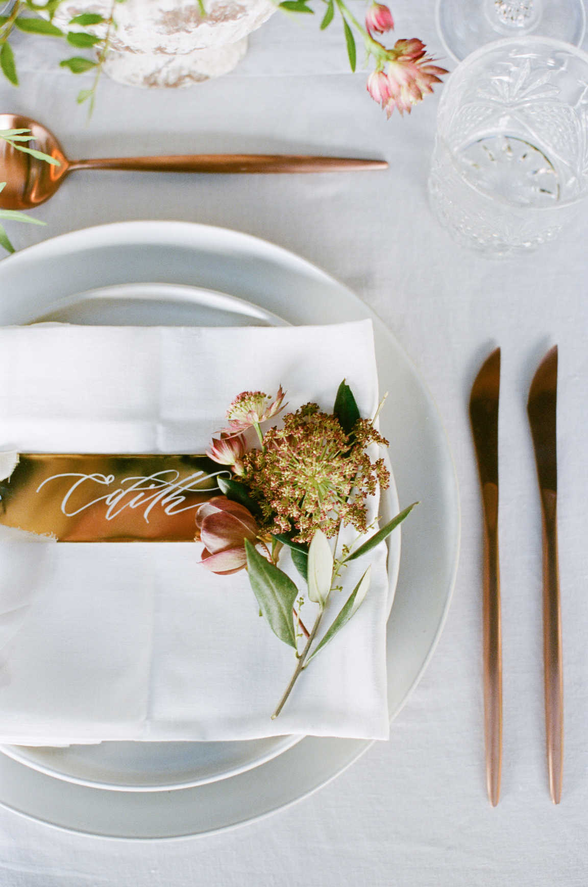 A top-down view of table setting with plates, knives, spoons, napkins, drink glasses, and a floral arrangement.