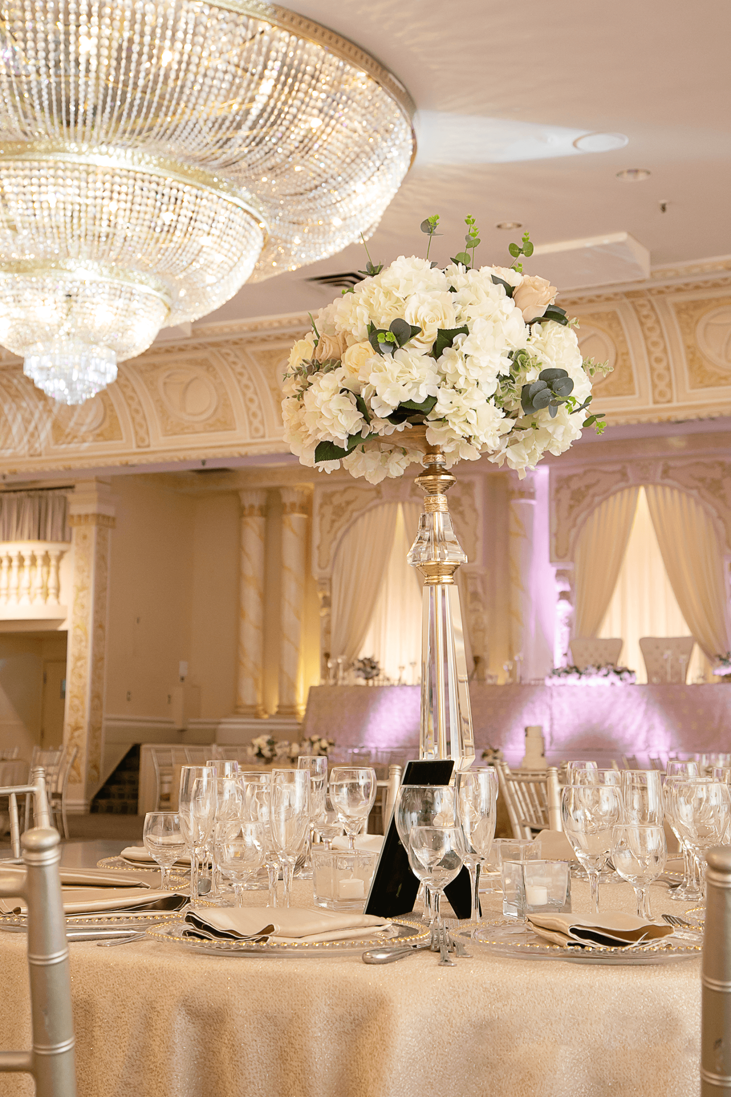 A bouquet of flowers in a vase atop a table at a wedding with a huge chandelier at the Grand Queens Wedding Venue at Paradise Banquet Halls.