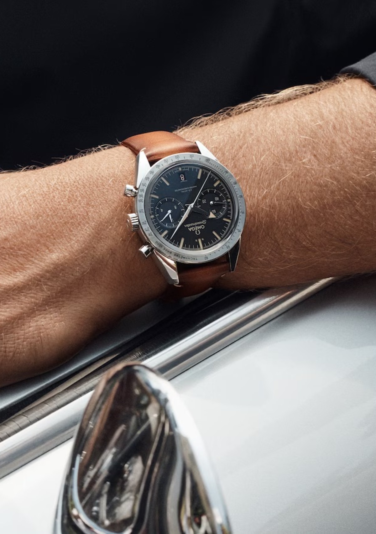 A close-up image of a person's wrist wearing a luxury chronograph watch with a brown leather strap. The watch face is black with multiple sub-dials and silver hour markers. The person's arm is resting on a car, with part of its chrome side mirror visible.