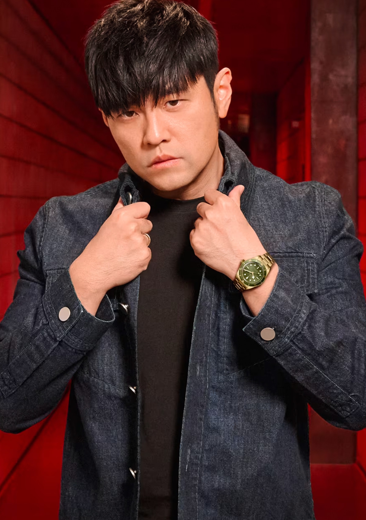 A person with straight, dark hair wearing a dark denim jacket and a gold watch stands in a dimly lit corridor with red walls, holding the collar of the jacket with both hands and looking directly at the camera.