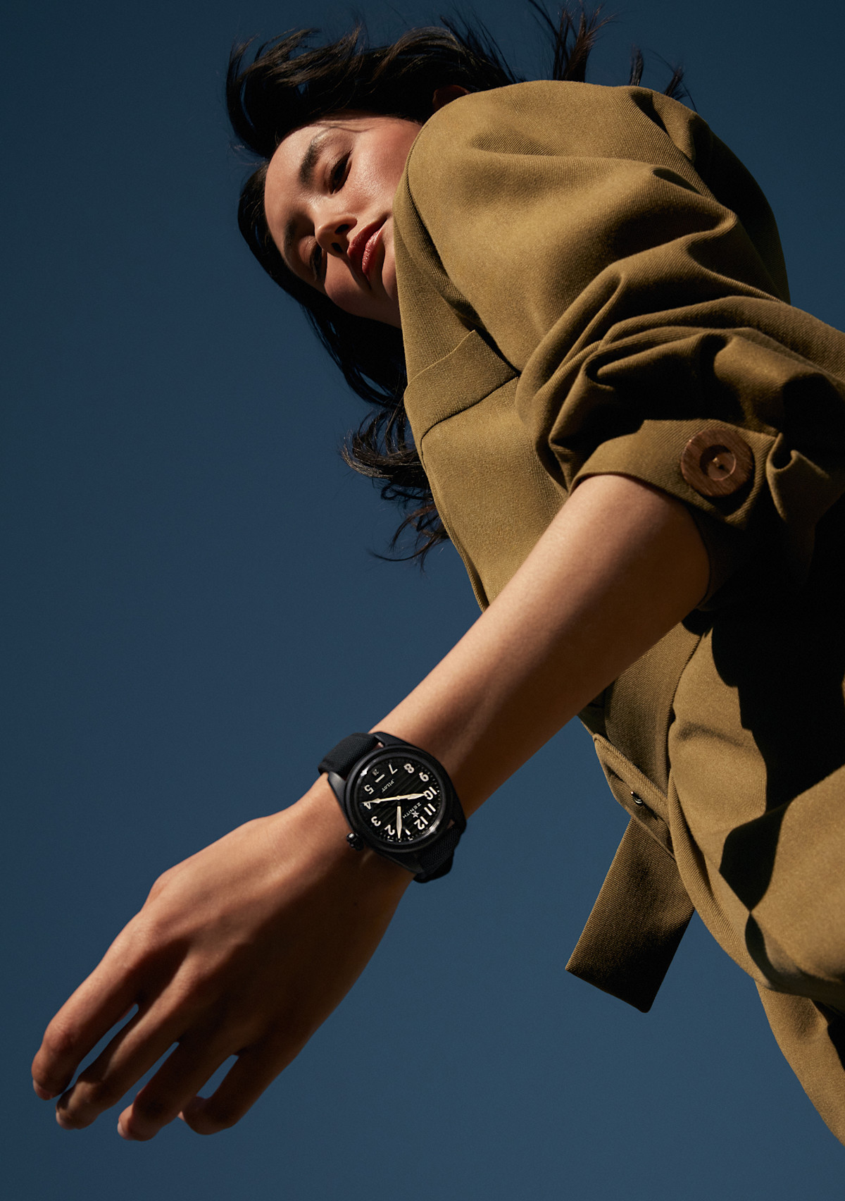 Low-angle view of a person wearing a tan jacket, seen from below against a dark blue sky. The person's wrist, adorned with a large black watch, is prominently in the foreground, positioned above their head, creating a dramatic and dynamic composition.
