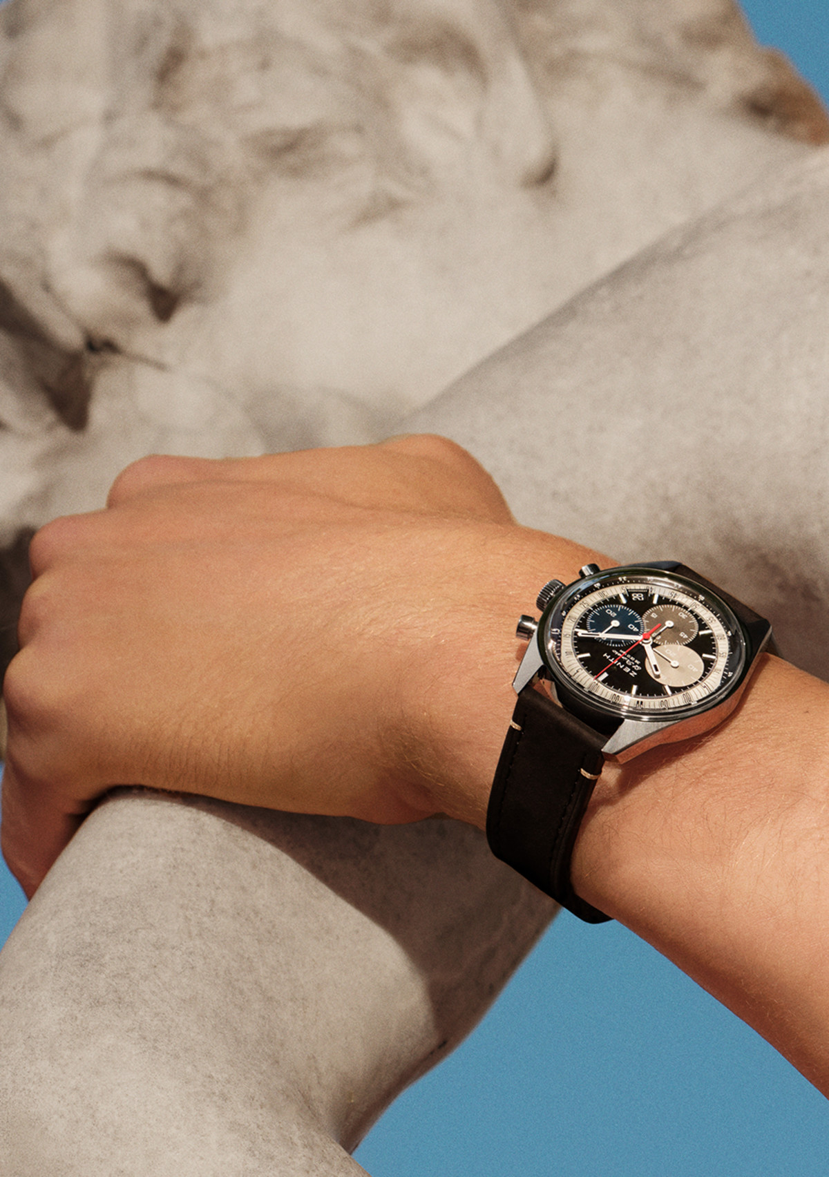 A person wearing a blue sweater with a white trim has their hand in the pocket of black pants. They are wearing a black analog wristwatch with a silver bezel and three subdials on their left wrist.