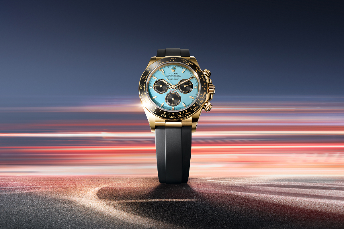 A luxury watch with a white and silver face, encrusted with diamonds around the bezel. The watch features three sub-dials with a mother of pearl design, and has a black leather strap. The background is a gradient of light blue and warm yellow hues.