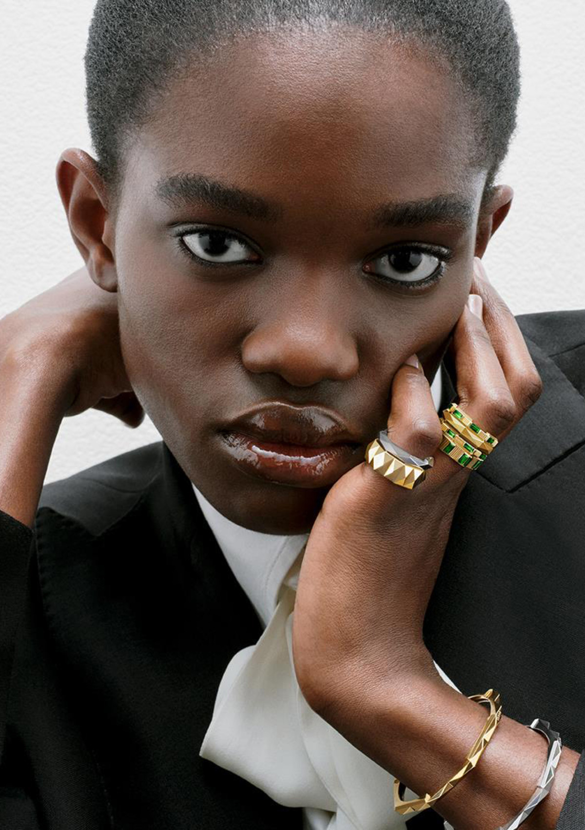 A person with short, dark hair gazes directly at the camera. They rest their face on one hand, fingers adorned with multiple shiny rings, while wearing a black jacket over a white top. The background is plain and light-colored.