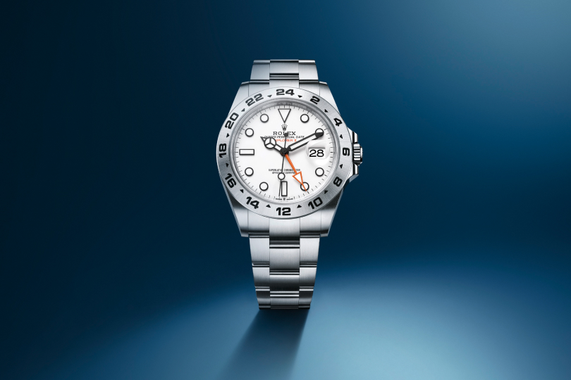 A luxury wristwatch with a two-tone metal band, featuring silver and gold links. The watch has a black face with gold hour markers, hands, and a prominent gold bezel. It is set against an abstract, gradient background with a rippling texture.