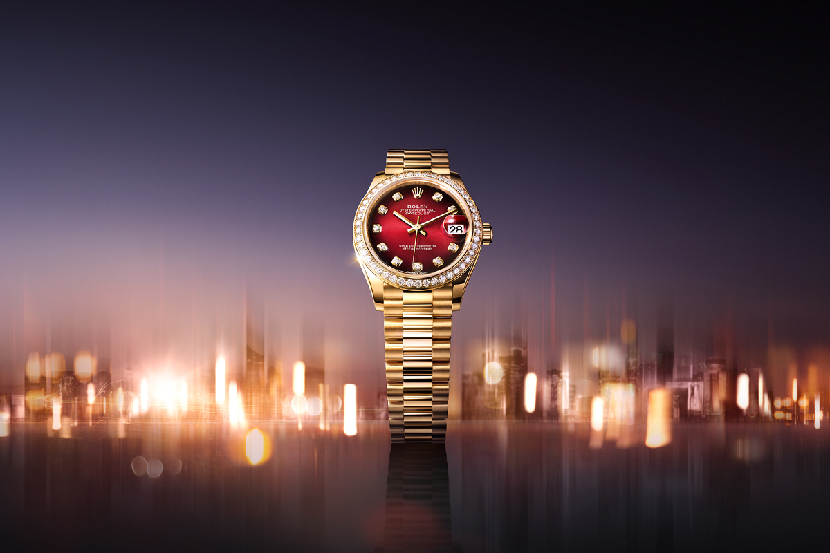 A luxurious rose gold watch with a metallic bracelet band against a blurred, gradient background. The watch has a black dial with rose gold hour markers and hands, as well as a date display. The watch is adorned with a fluted bezel and prominently displayed crown logo.