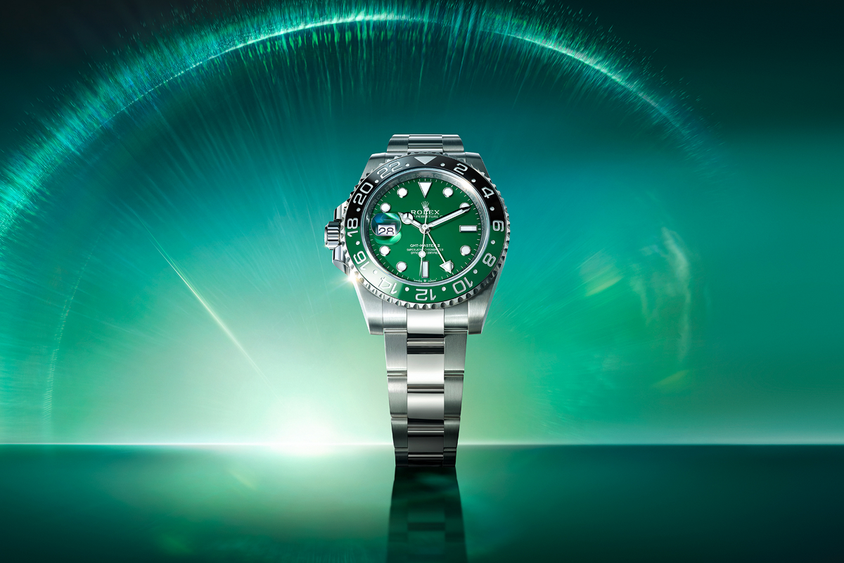 A stainless steel wristwatch with a dark face, luminous hands, and a date display at the 3 o'clock position. The bezel is marked with 24-hour increments. The background is gradient shades of blue and green. The watch is displayed in an upright position.