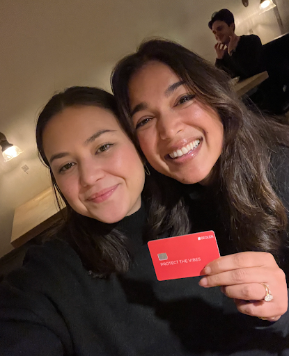Two young women holding up an orange Sequin Visa® Debit Card with writing on the card saying "Protect the Vibes"