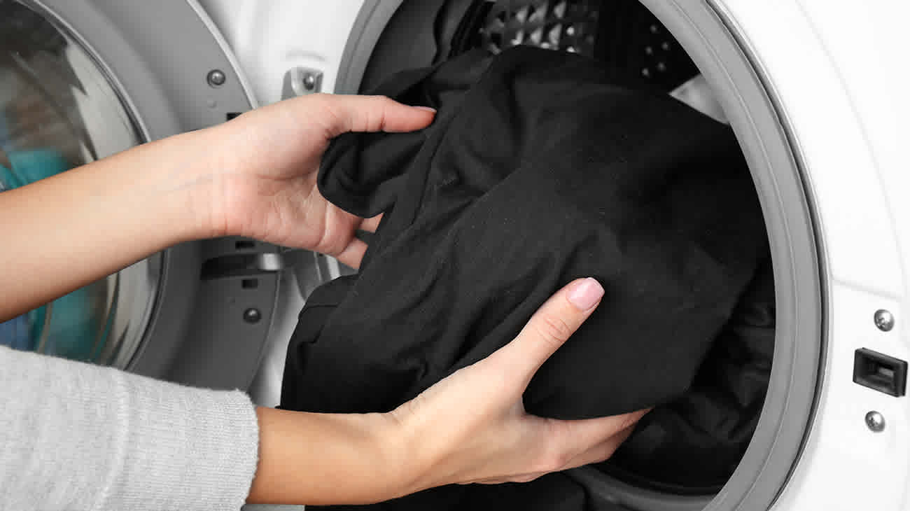 Manos femeninas acercando ropa oscura al lavarropas.