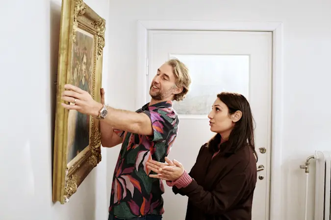 En man och en kvinna hänger upp en stor inramad tavla på en vit vägg i ett hem, nära en dörr och ett element.