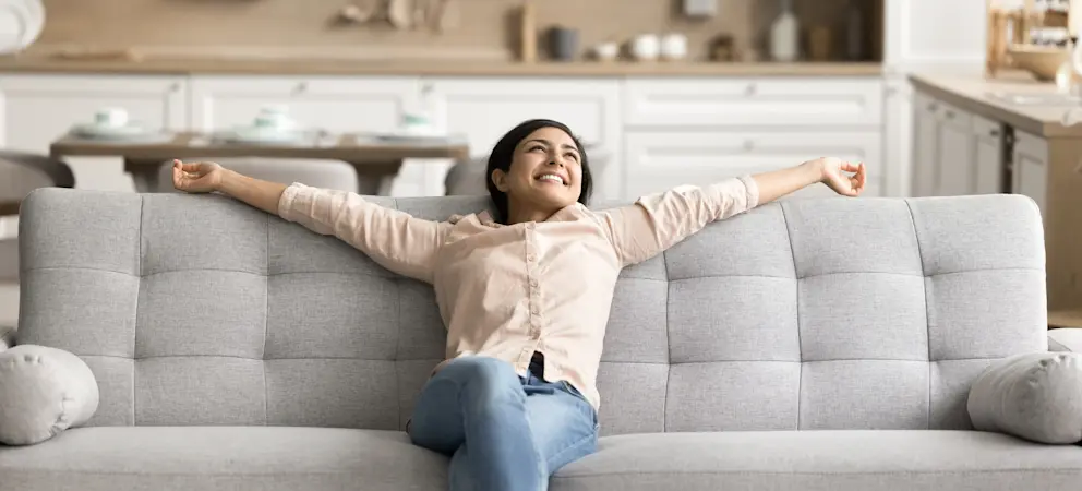Person sitter utsträckt i grå soffa i vardagsrum med öppet kök i bakgrunden och ljus, modern inredning.