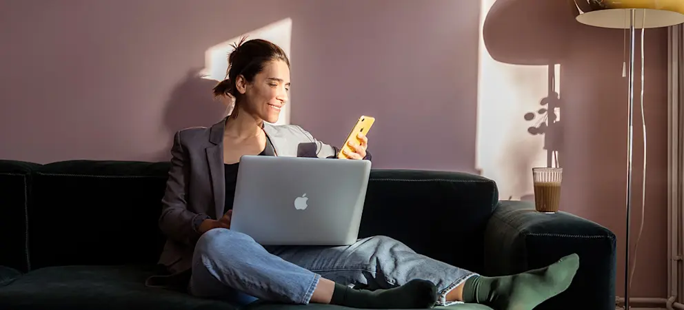 Person sitter i solbelyst vardagsrum på mörk soffa med laptop och mobil bredvid en golvlampa.