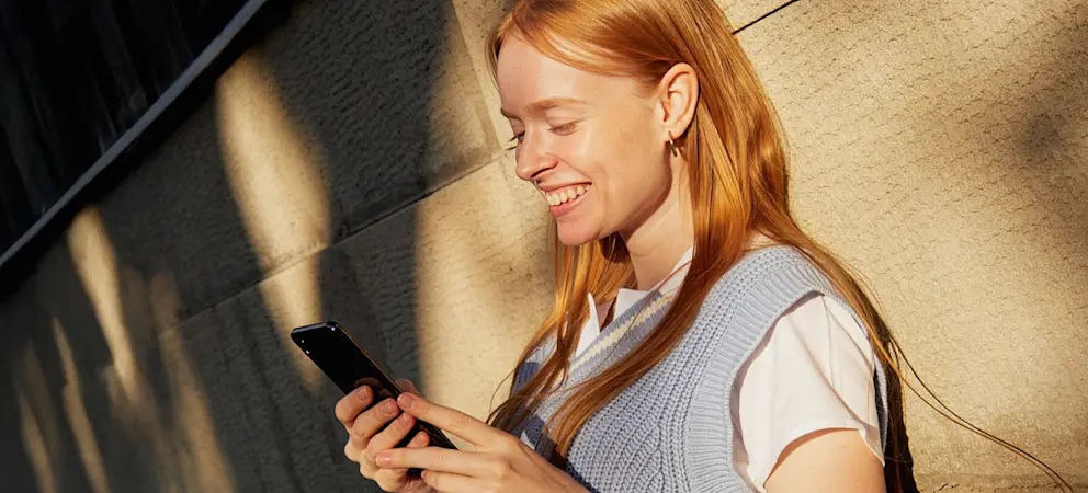 Person använder mobiltelefon utomhus i kvällssol mot byggnadsfasad i modern stadsmiljö.