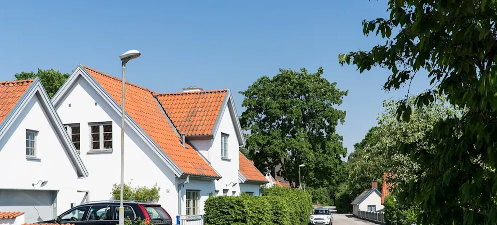 Villor med vita fasader och tegeltak längs lugn gata med grönska och parkerade bilar i bostadsområde.