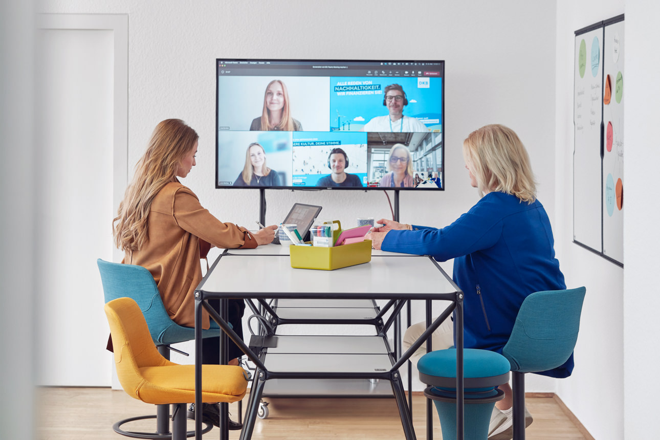 Zwei Frauen sitzen vor einem großen Monitor und nehmen an einer Videokonferenz teil