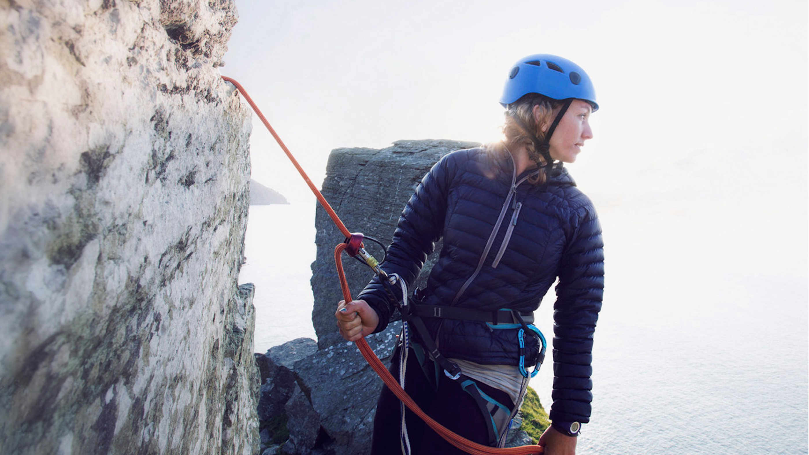 Eine Bergsteigerin, welche an einem Felsen abgesichert ist macht eine Rast und blickt über die Schulter.