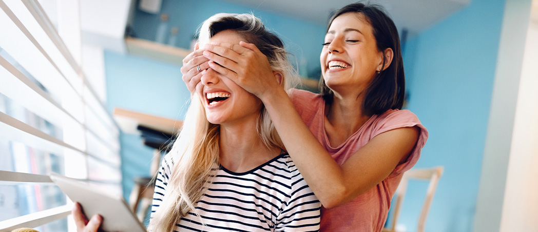 Eine Frau hält einer anderen Frau die Augen zu während sie lachend vor einem Tablet sitzen