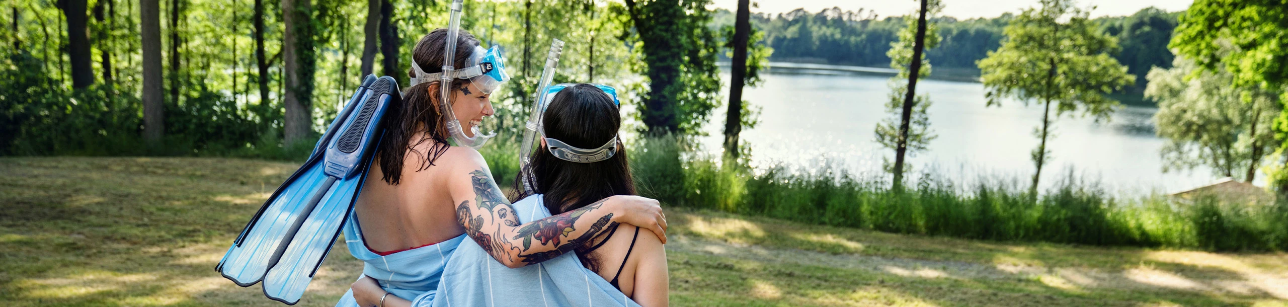 Zwei Personen mit Taucherausrüstung auf grüner Wiese am See