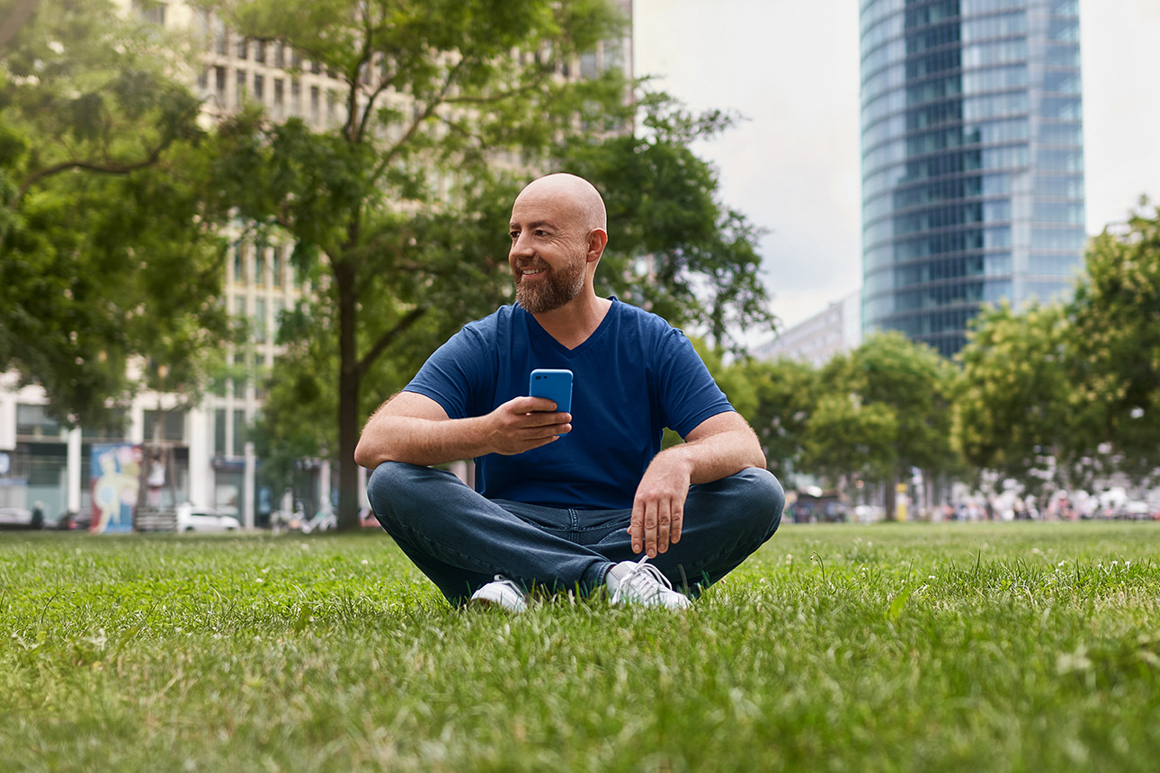 Ein Mann sitzt auf einer Wiese und hält ein Handy in der Hand.