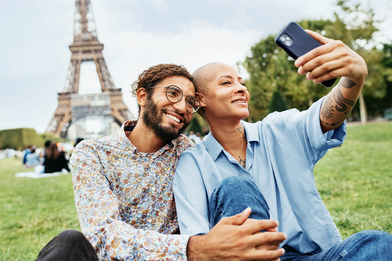 Bildbeschreibung: Ein Pärchen sitzt vor dem Eifelturm in Paris und macht ein Selfie.
