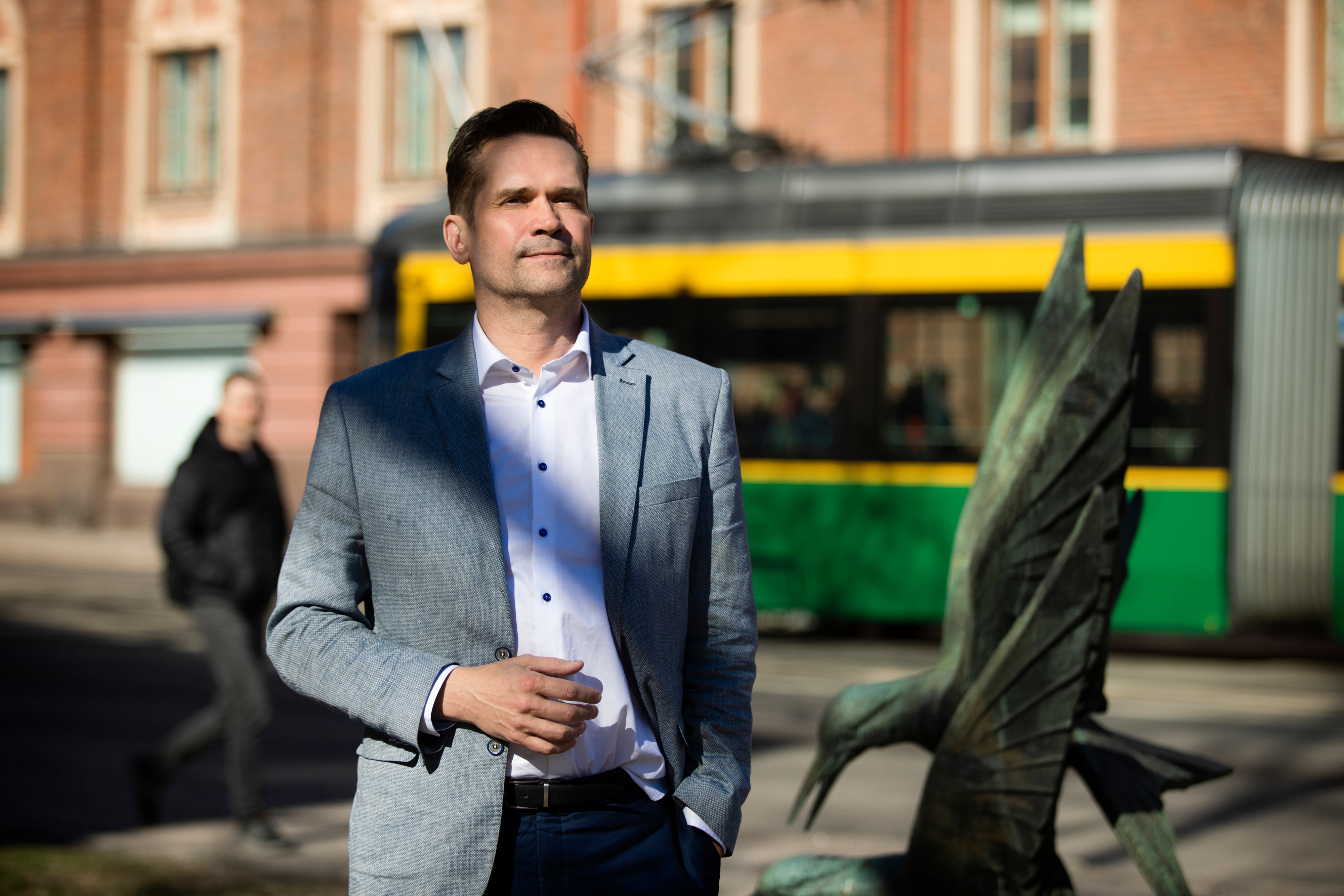Mika Aaltola seisoo ulkona kadulla lintupatsaan edessä, taustalla kulkee raitiovaunu.