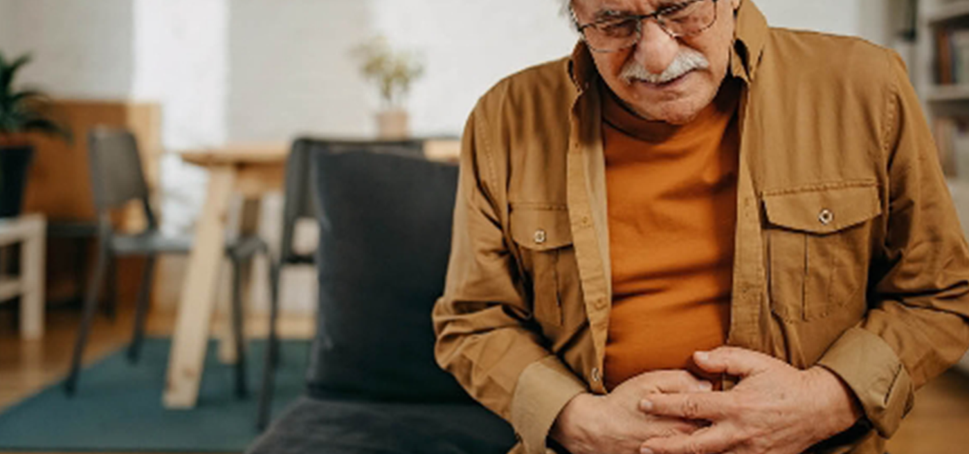 imagen que muestra a un señor de mediana edad sentado en un sofá apretándose con las manos en la zona del abdomen y con cara de dolor