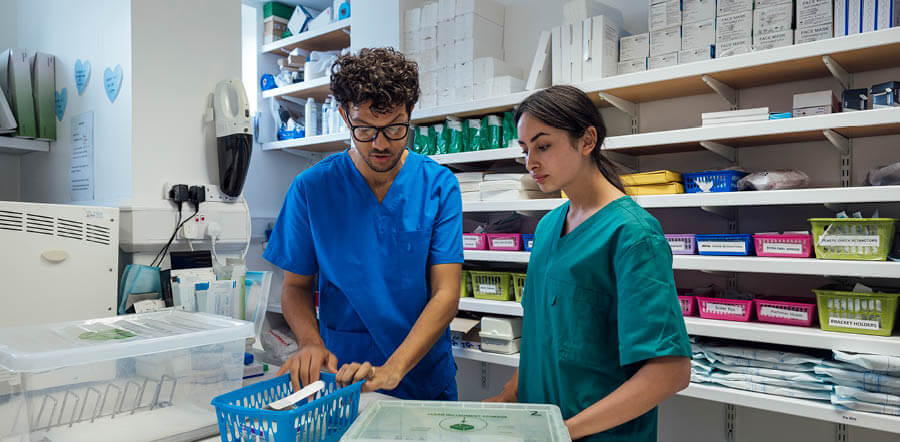 La imagen muestra a dos profesionales sanitarios en una sala mirando a una caja con medicamentos que se situa encima de un escritorio