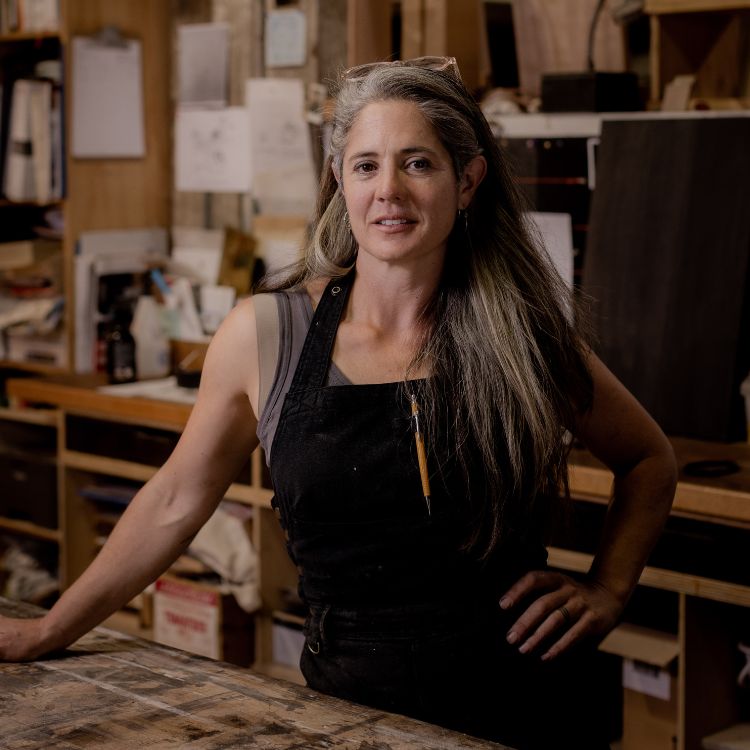 A woman with long silvering hair wearing an apron with a pen hanging from the centre, leaning on a bench with tools and paperwork behind her