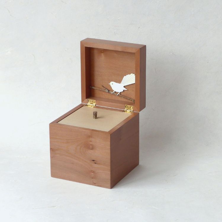 A small box, a hinged lid open to reveal a second lid with handle to open. A small white bird is depicted, perched on a branch, on the inside of the hinged lid.
