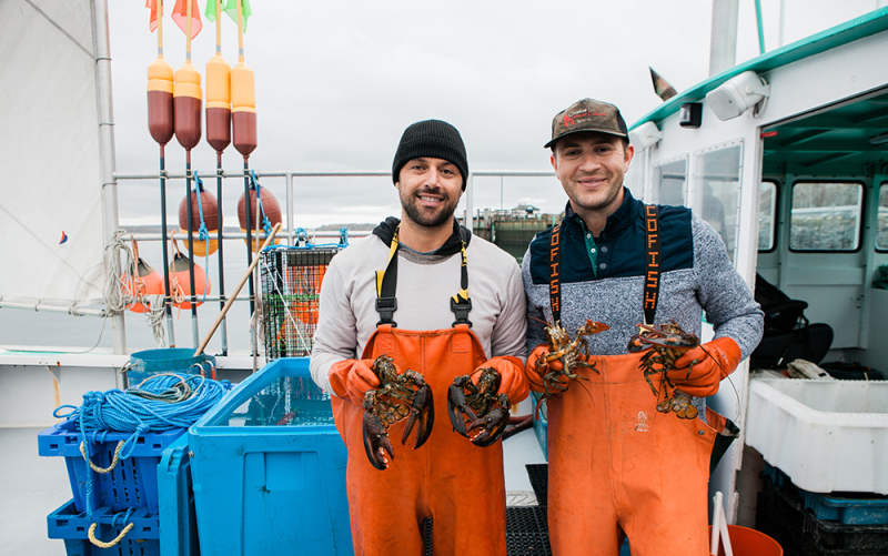 Cousins Maine Lobster founders Jim and Sabin are on a fishing boat, holding live Maine lobsters.