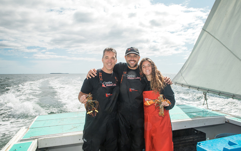 A photo of Cousin Sabin on the back of a lobster boat, with his arms around two members of our Boston family, as they both hold live lobsters.