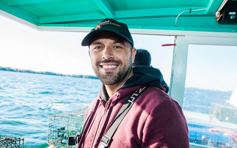 A closeup photo of Cousin Sabin smiling in a hoodie and a CML hat on board a lobster boat in Maine.